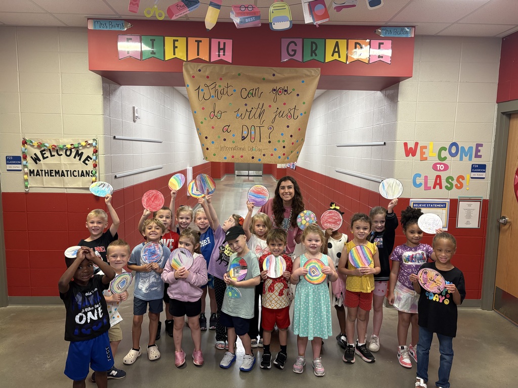 Class stanign together holding circles they colored under a sign in the hallways that says "what can you do with just a dot?"