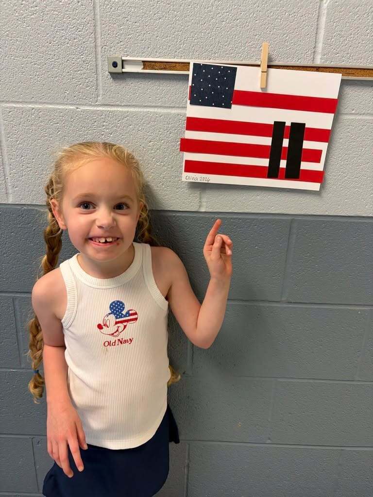 A child smiles for a photo in a school hallway and points up to a piece of American flag artwork.