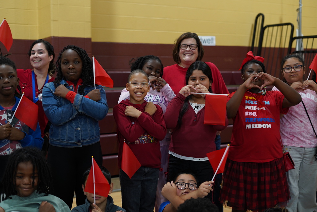 Students and teachers smile for a photo in a school gym. They hold up red flags.