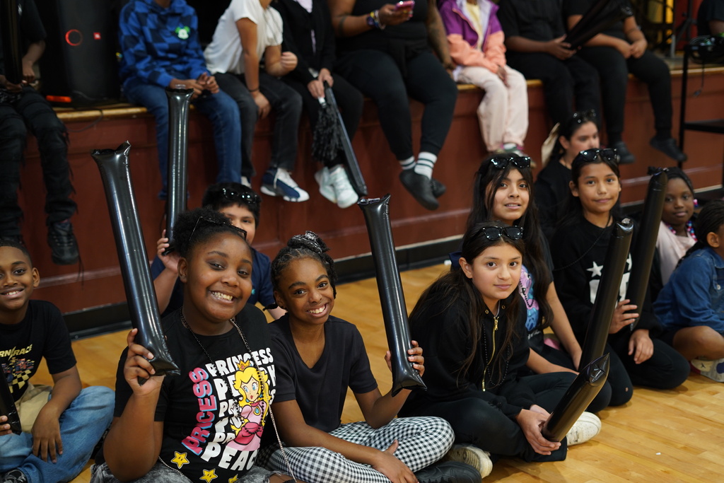 Students smile and hold up noise makers while sitting in a school gym.