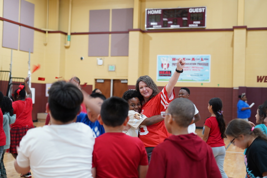 A student and a teacher hug in a crowded school gym.