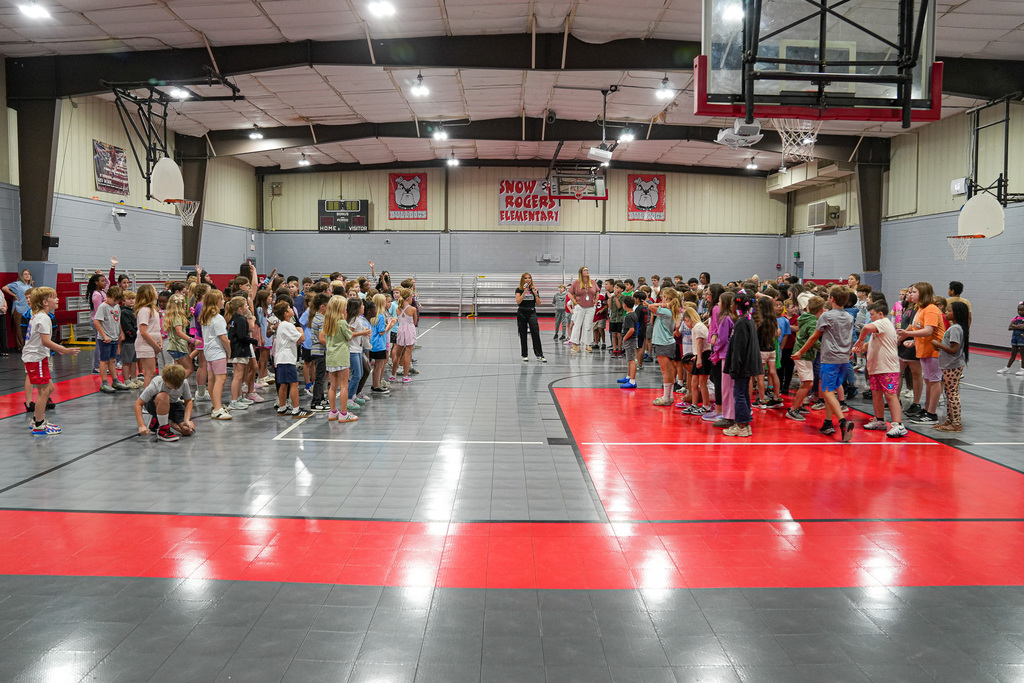 A woman speaks to a group of students while they stand inside a school gym.