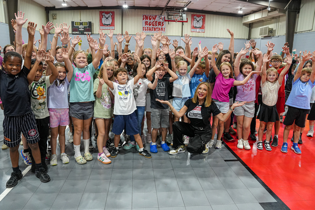 Students smile and hold their hands in the air while posing for a photo with a woman in a school gym.