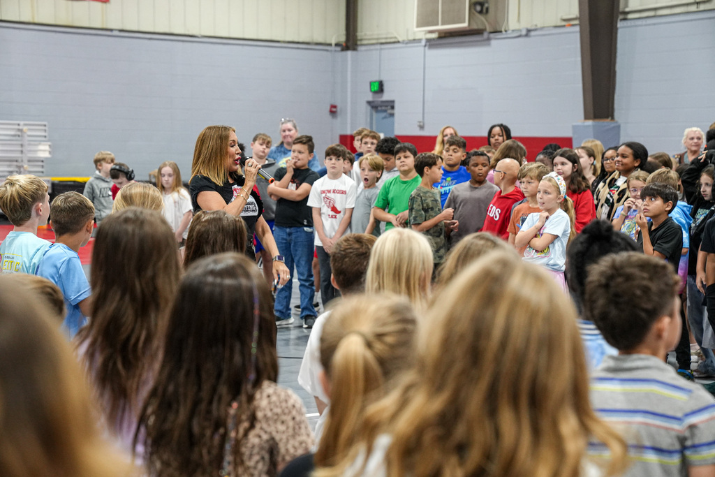 A woman speaks to students while they stand inside a school gym.