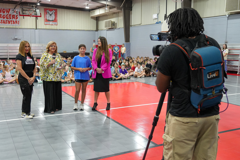 A camera person videos a newscaster interviewing women and a student in a school gym.