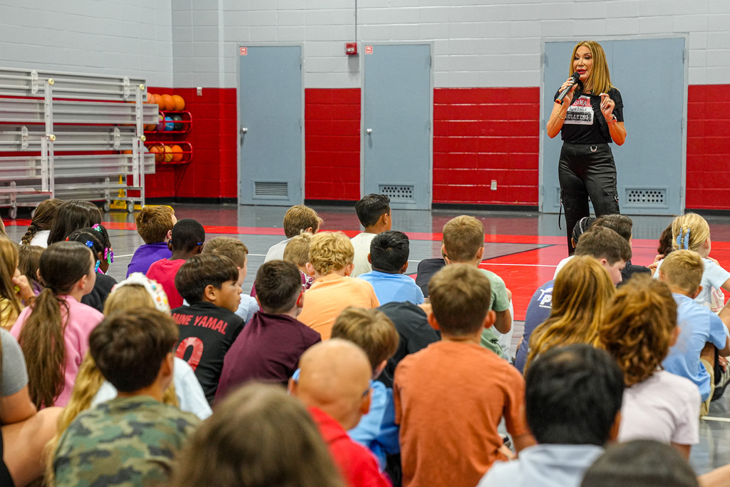 A woman speaks into a microphone while standing in front of a group of students in a school gym.