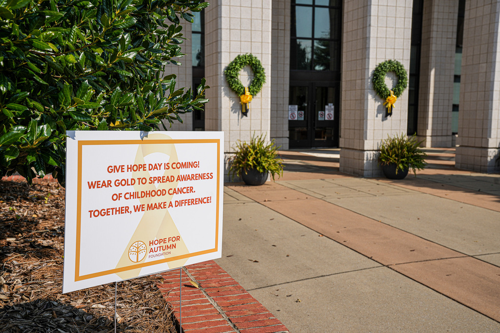 Sign advertising Give Hope Day is displayed outside a school. In the background, wreaths with yellow ribbons on them hang on the school building.