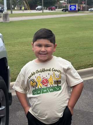 A child smiles outdoors for a photo while wearing a Childhood Cancer Awareness shirt.
