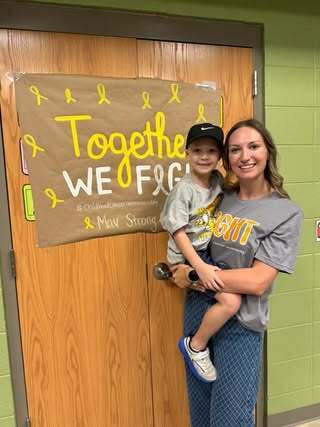 A woman smiles while holding a child. They stand in front of a sign for Childhood Cancer Awareness Month.