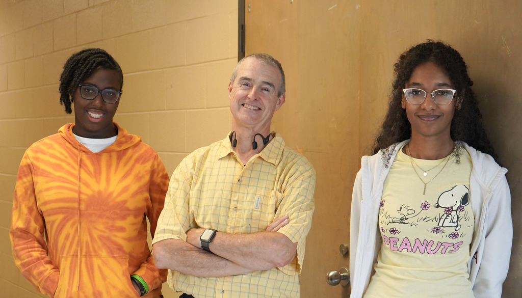 Students and a teacher smile for a photo in a school hallway. They all wear yellow shirts.