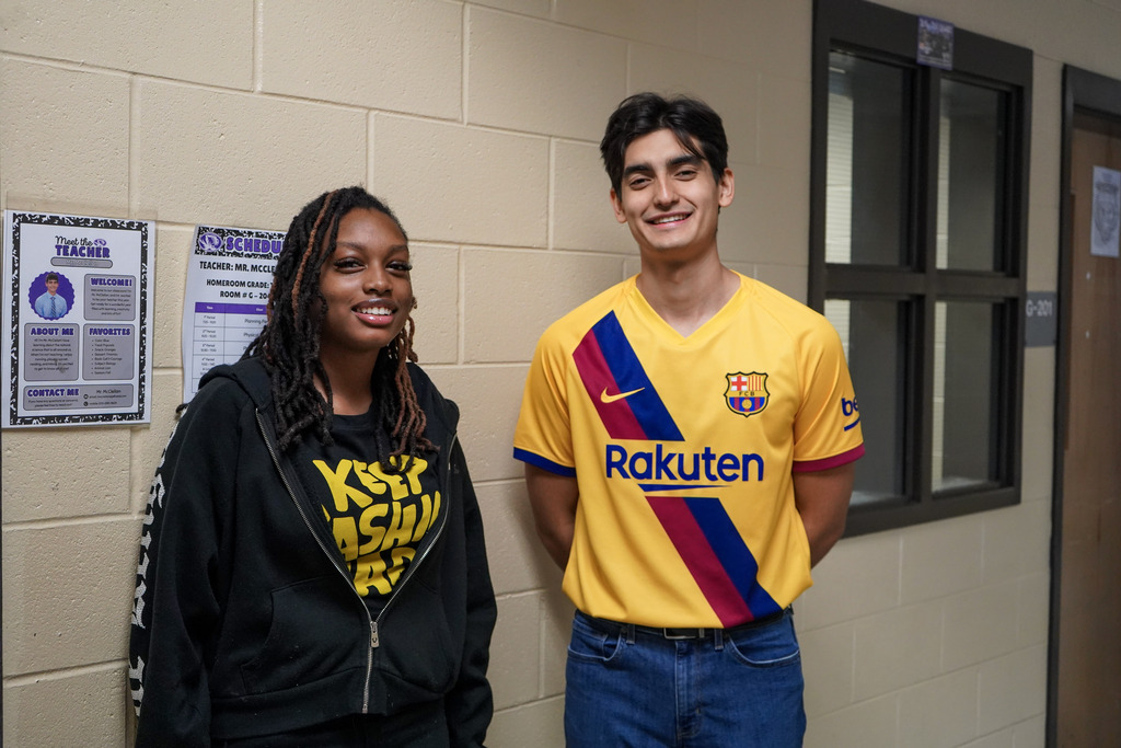 A student and a teacher smile for a photo while standing in a school hallway. They both wear yellow.