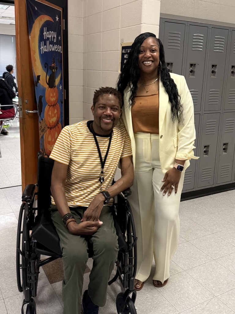 A man and a woman smile for a photo in a school hallway. They both wear yellow.