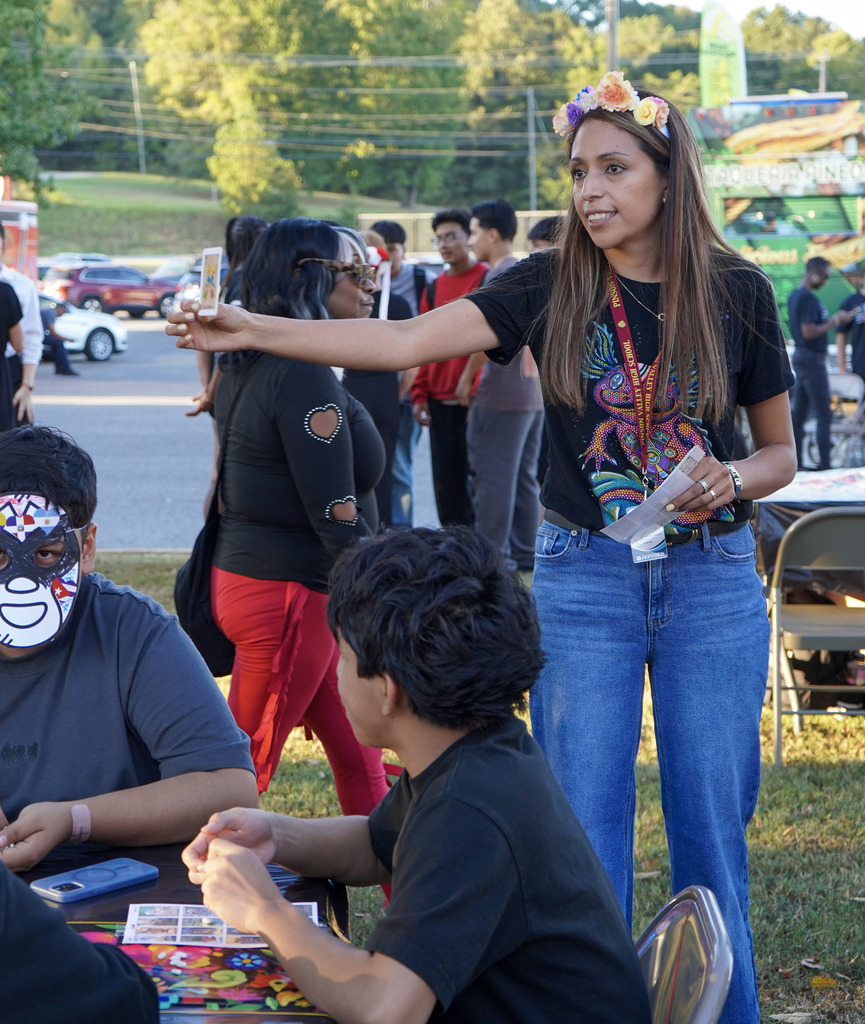 A woman smiles and holds up a card while young people sit at tables around her playing a game.