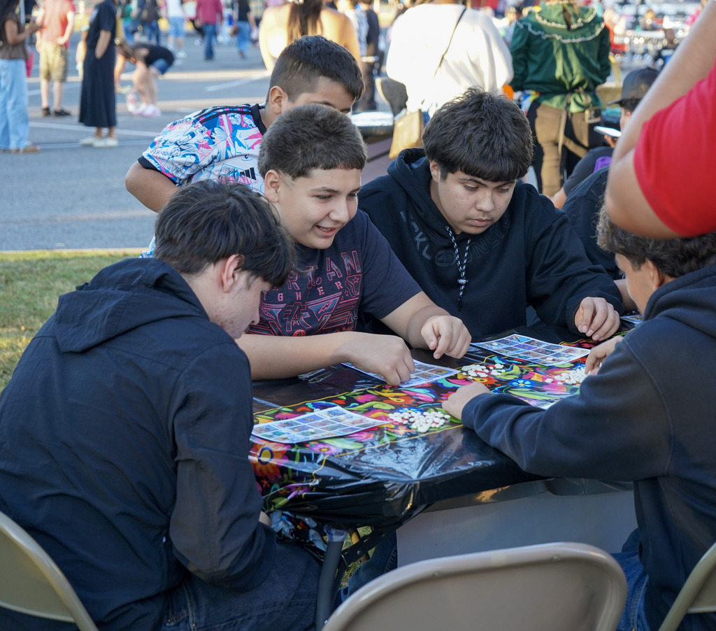 Young people play a board game while sitting outside at tables.