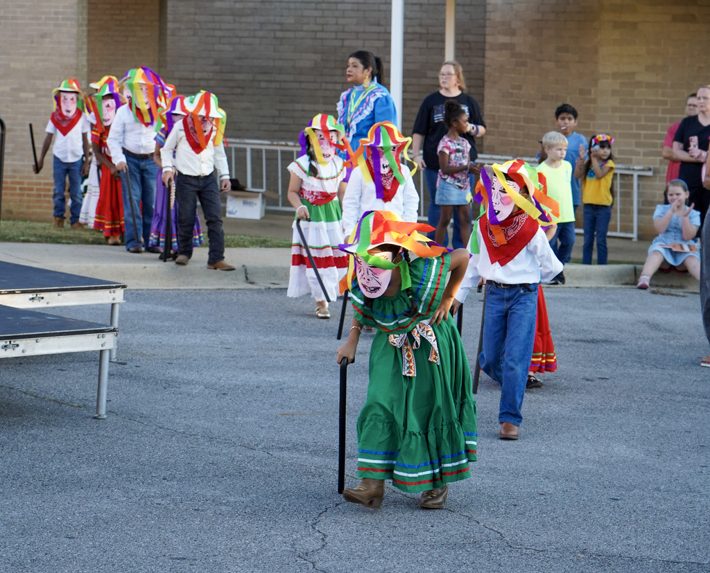 Children wearing bright hats, masks and costumes walk out into a parking lot for a performance during a festival.
