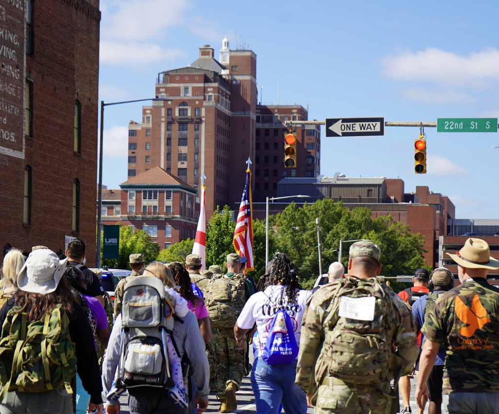 People walk down a street wearing weighted backpacks during a memorial ruck honoring veterans lost to suicide.
