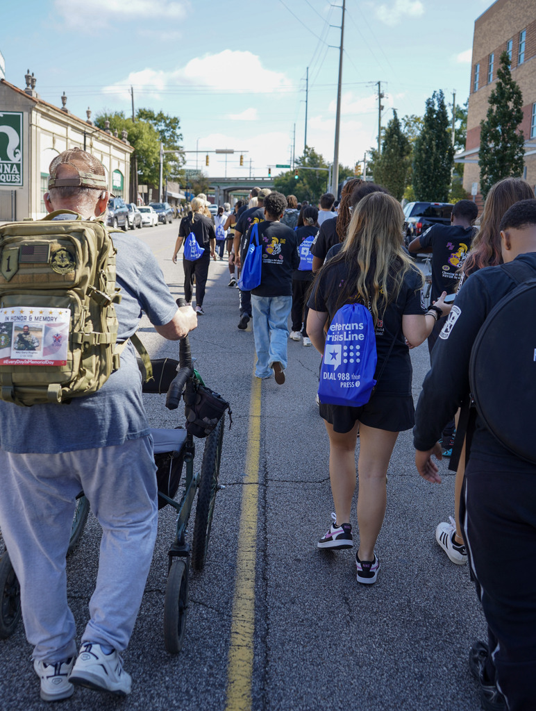 Community members and students walk down a street wearing weighted backpacks to honor veterans lost to suicide.