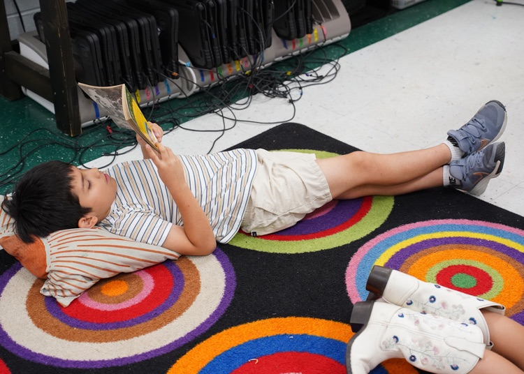 A student lays on top of a colorful rug and reads a book held above his head.