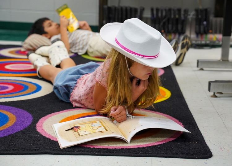 an elementary school student dressed as Dolly Parton lays on the floor and reads a book.
