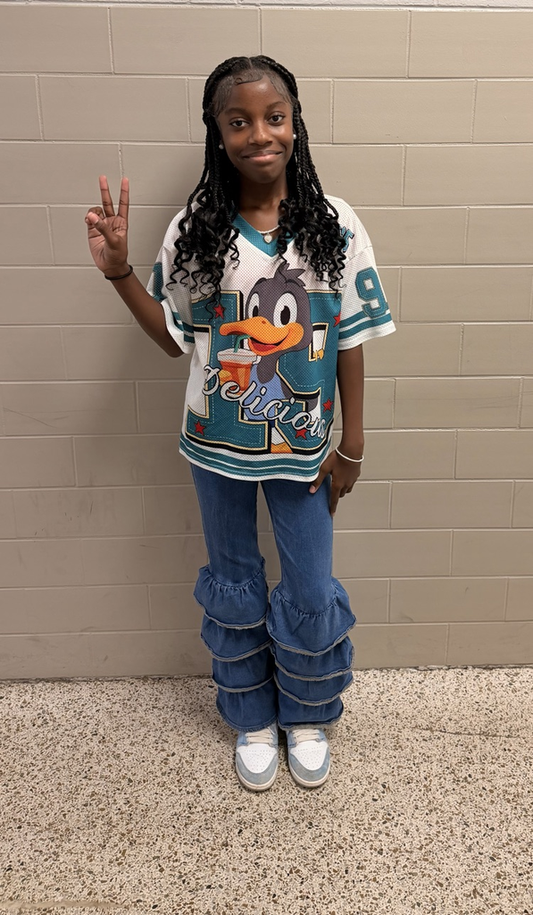 HMS student smiles and gives a peace sign while wearing a colorful sports-style jersey and jeans.
