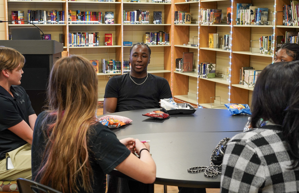 Students sit at a table in a library and speak to each other.