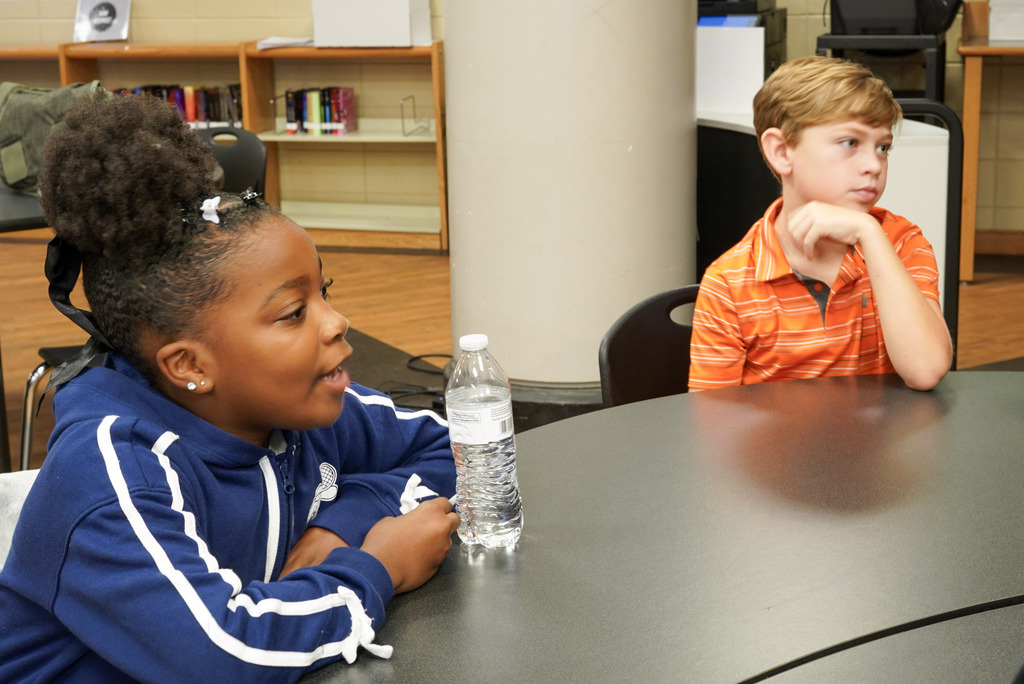 Children sit at a table inside a school library. One of the children is talking.
