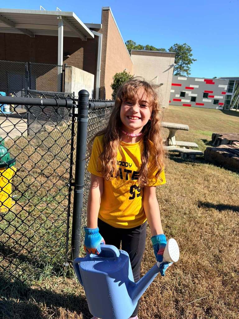 A student smiles while holding gardening materials outdoors.