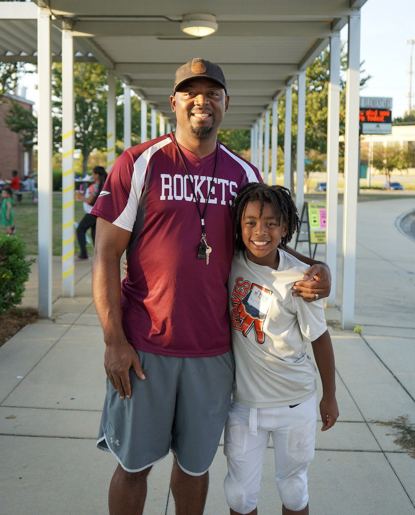 A man and a child hug and smile for a photo while standing outside a school gym.