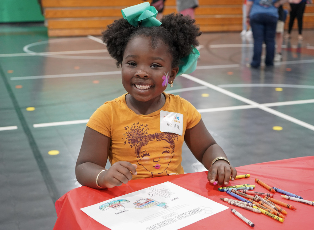 A child smiles while coloring on a piece of paper inside a school gym.