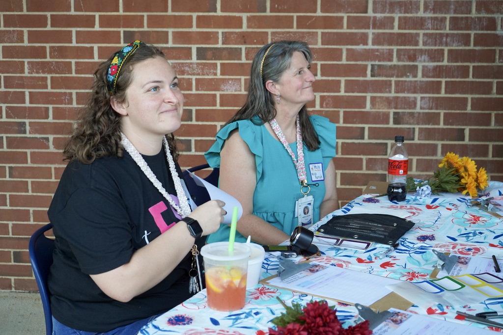 Two women smile while sitting at a table outside a school.
