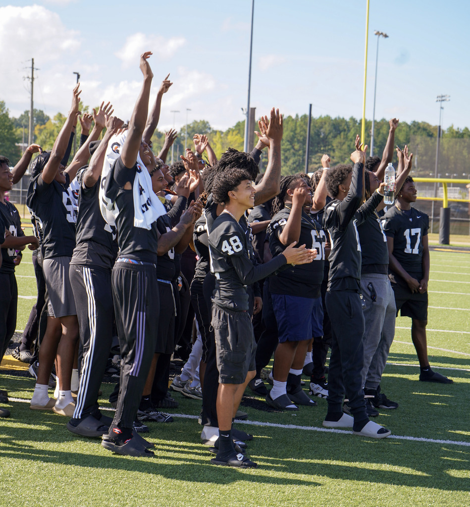 Football players hold their hands in the air and cheer while standing on a football field during a pep rally.
