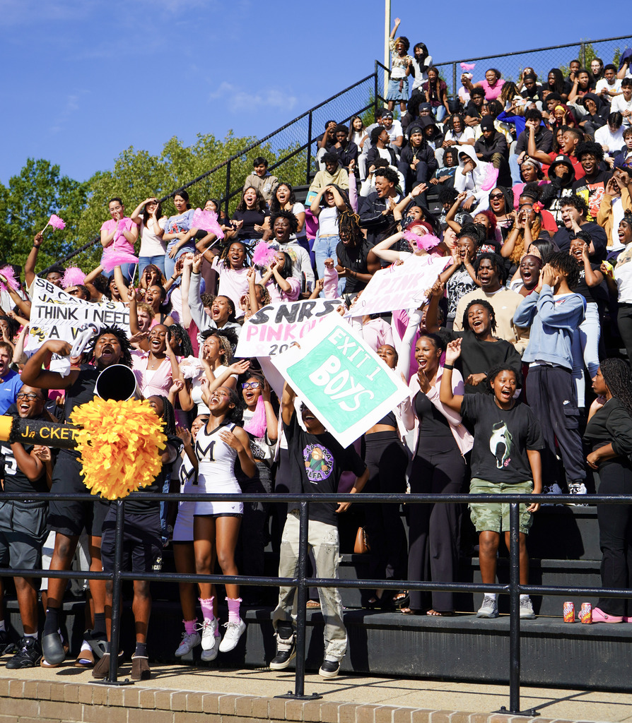 Students stand in bleachers and cheer while holding signs.