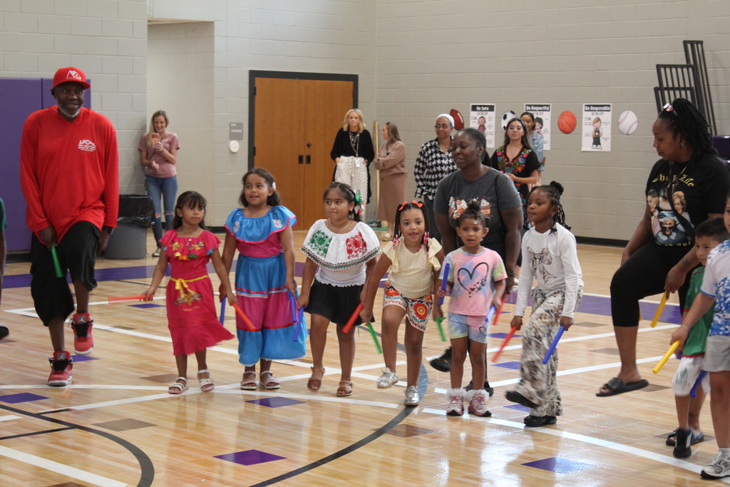 Students in a circle in the gym holding rhythm sticks while performing a dance