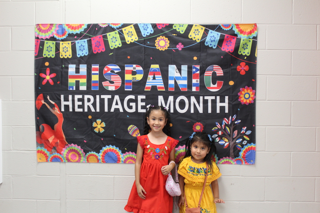Two students in traditional hispanic dresses in front of a sign that says Hispanic Heritage Month