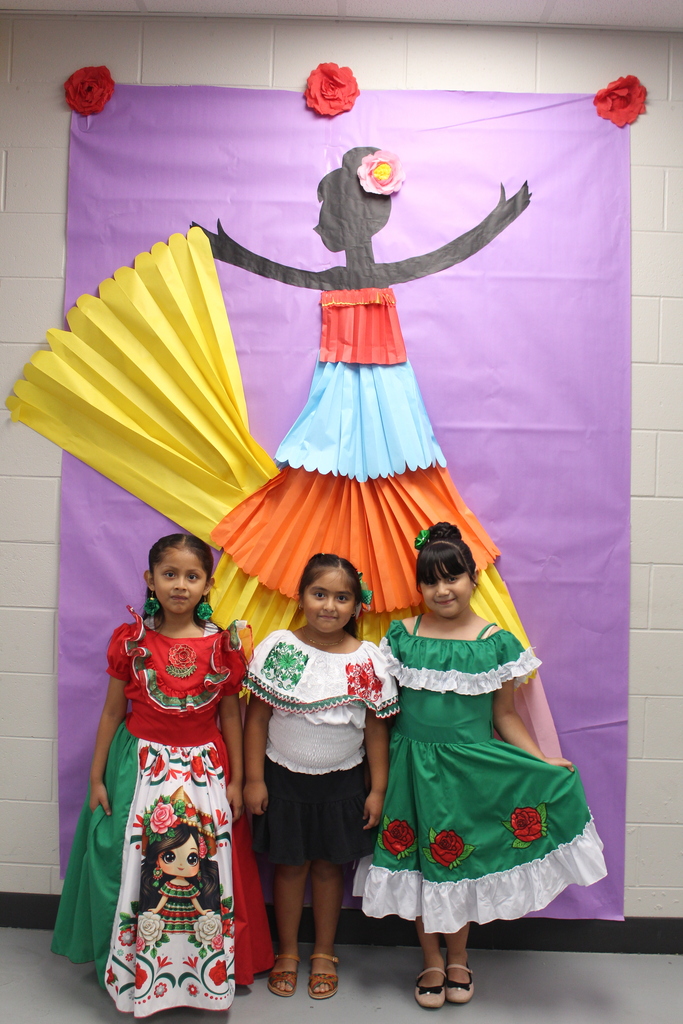 Three students in traditional hispanic dress in front of a mural of a girl in a hispanic dress