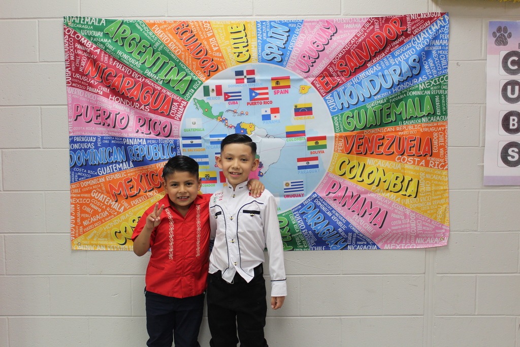 Two students in traditional hispanic clothing in front of a flag with a map and all of the names of spanish speaking countries