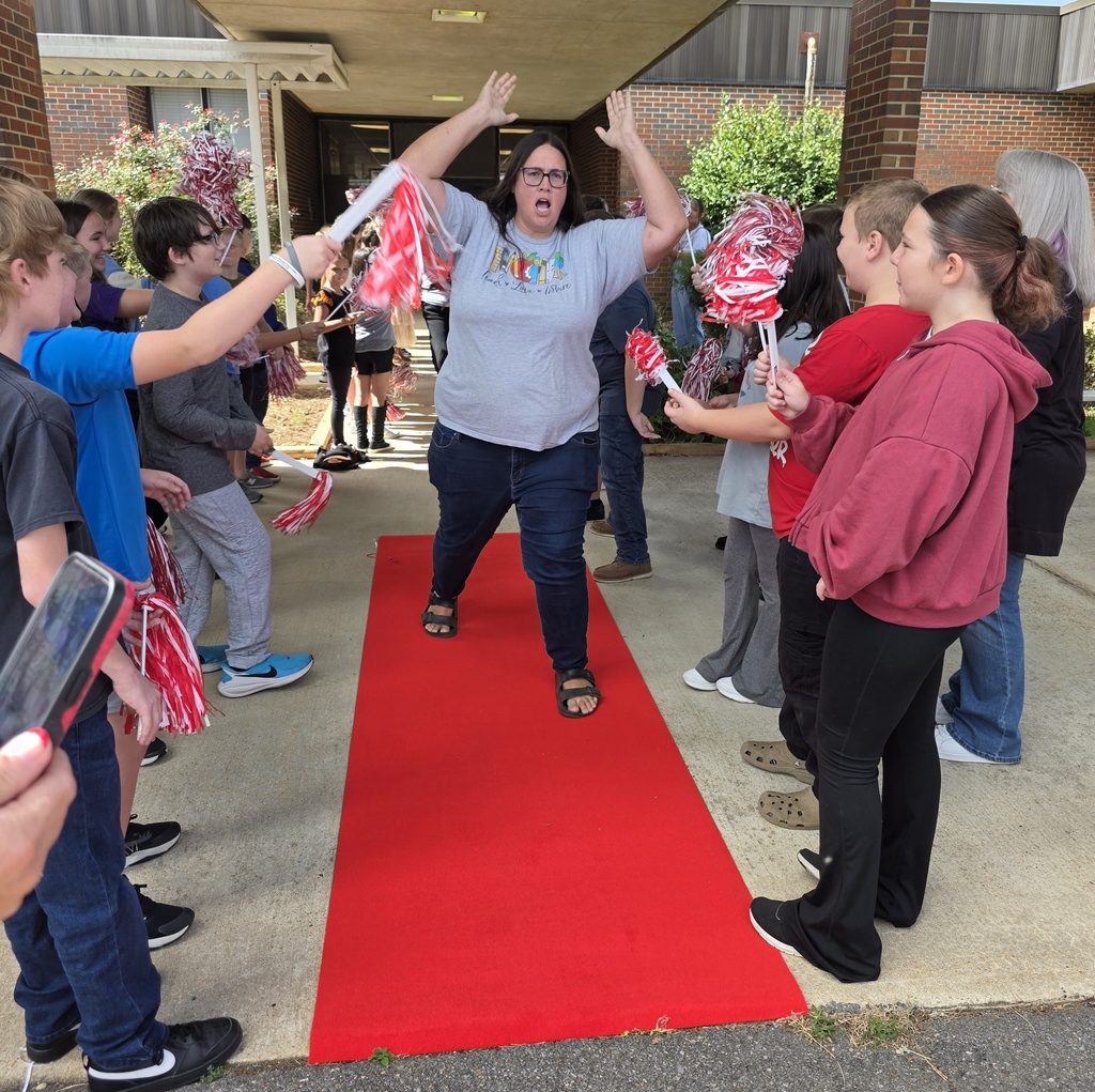 teacher walking down a red carpet with students shaking shakers and cheering her on