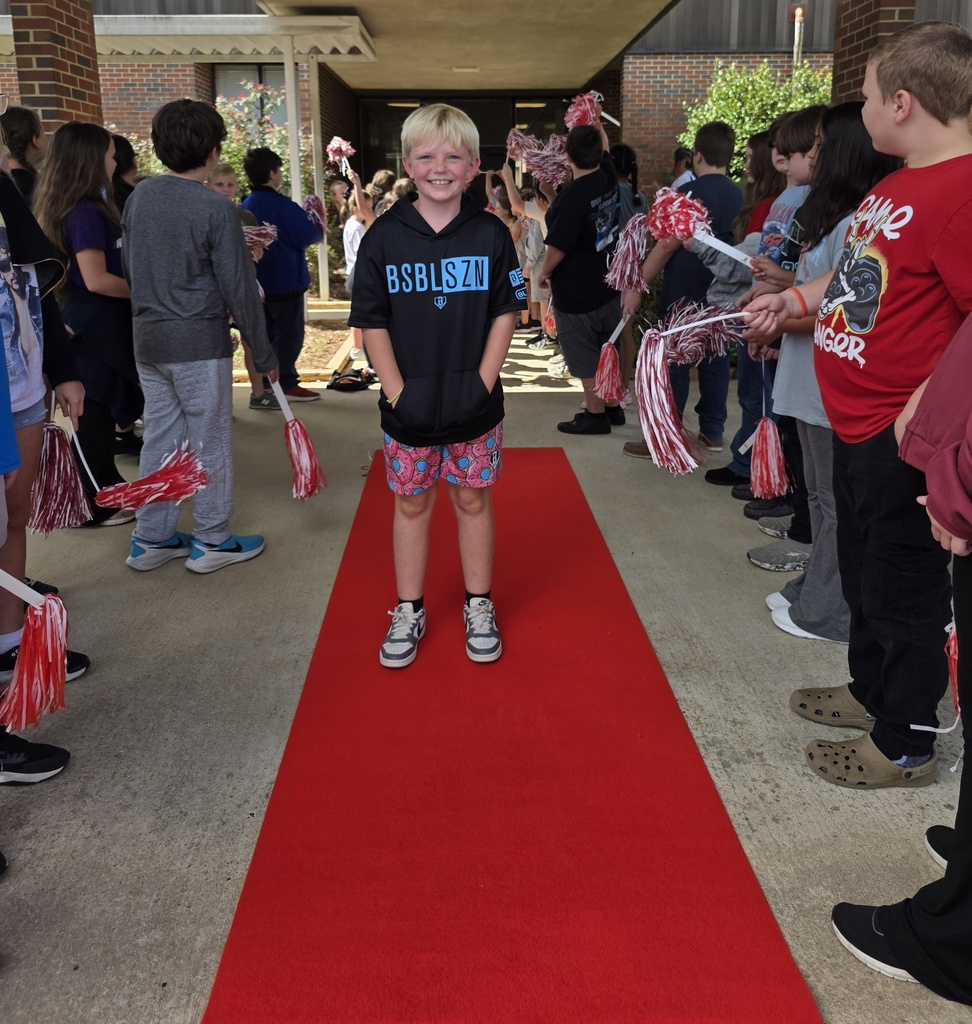 boy standing on red carpet with students shaking shakers around her