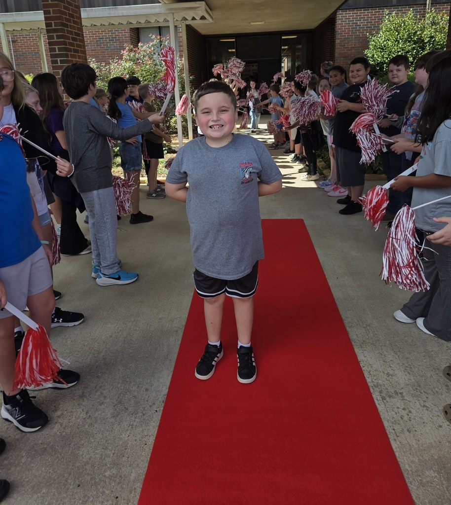 boy standing on red carpet with students shaking shakers around her