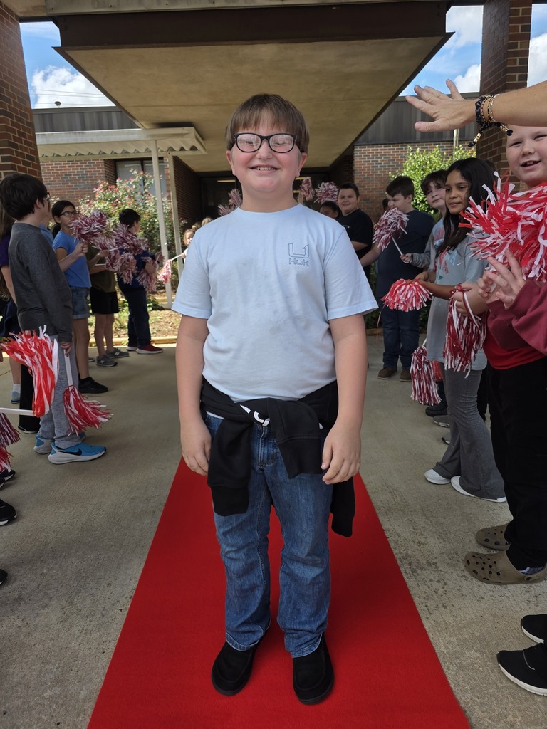 boy standing on a red carpet with students shaking shakers near him