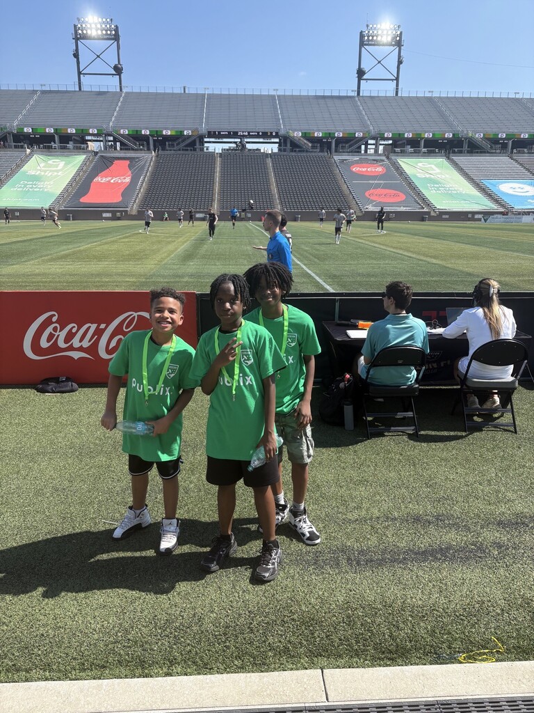 Three McAdory students in green shirts smiling on the sideline during the Birmingham Legion FC game.