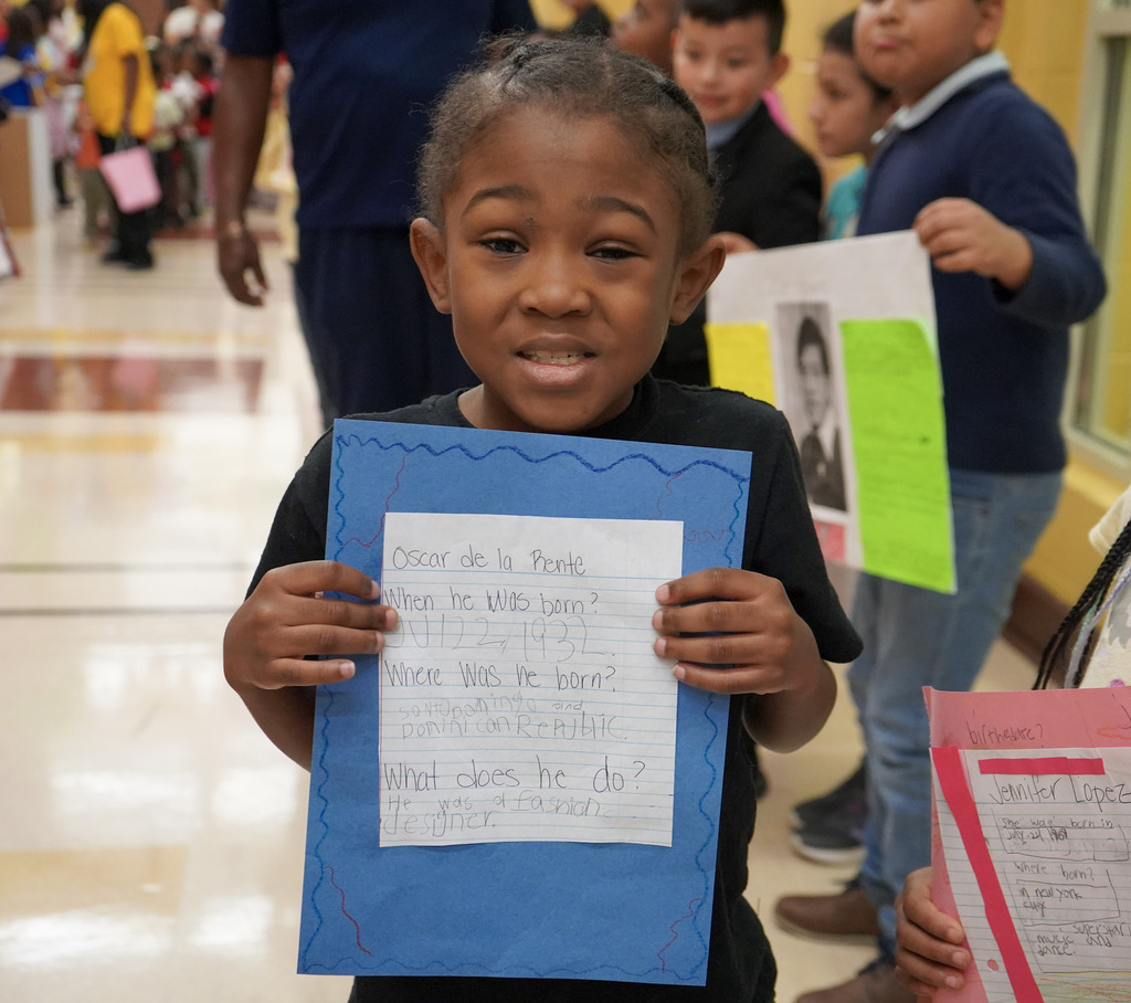 A child smiles while holding up a piece of paper and standing inside a school hallway. The paper has facts about Oscar de la Renta.