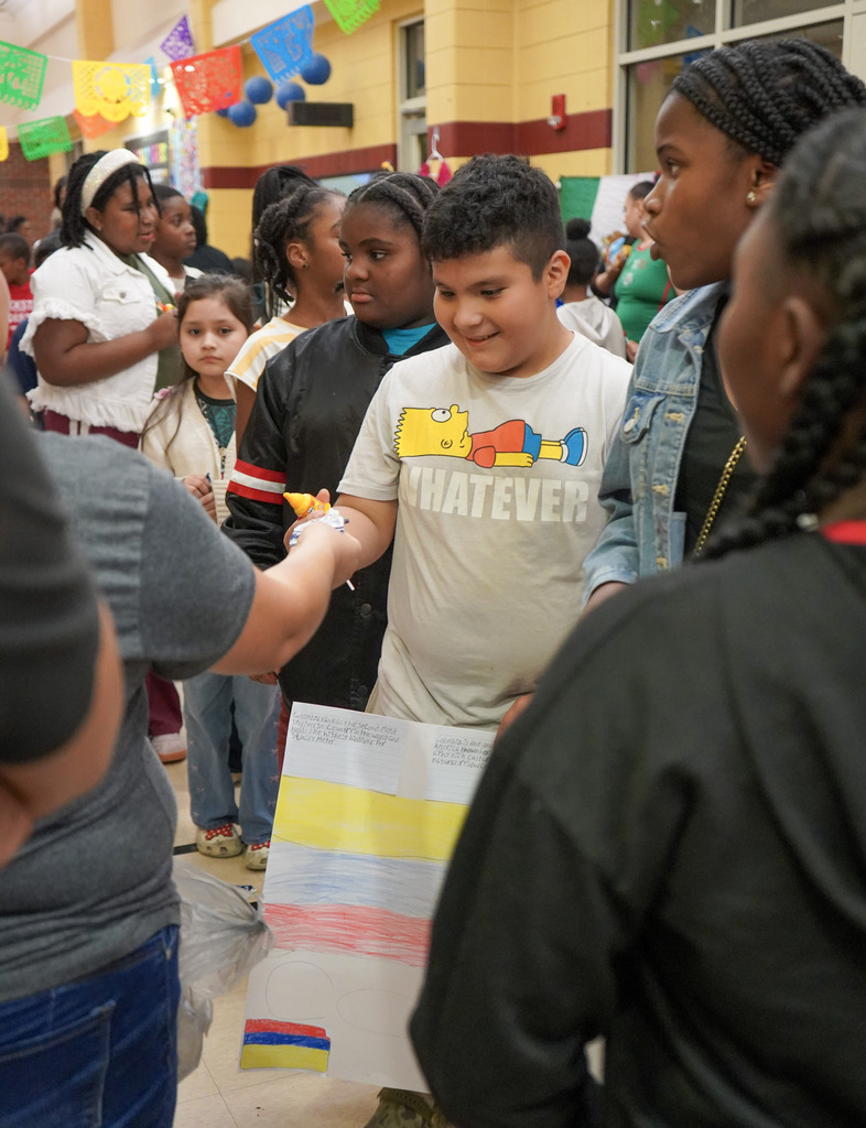 An adult hands a child a piece of candy during a Hispanic Heritage Festival inside a school hallway.