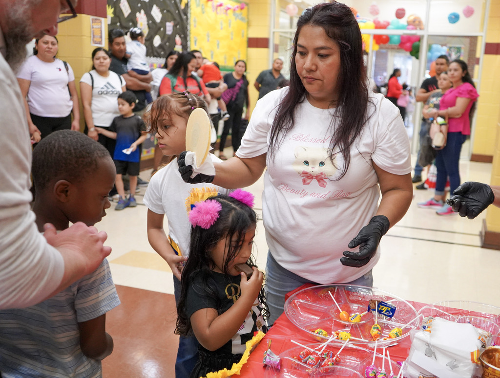 A woman hands tortillas to a student as he stands inside a school hallway. Children and families stand around them.