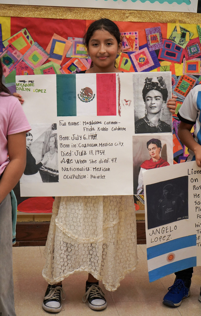 A girl holds a hand-made poster featuring facts about Frida Kahlo while standing in a school hallway.