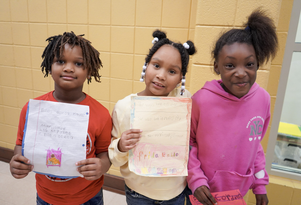 Three children stand together and smile for a photo inside a school hallway. They hold pieces of paper if their hands.