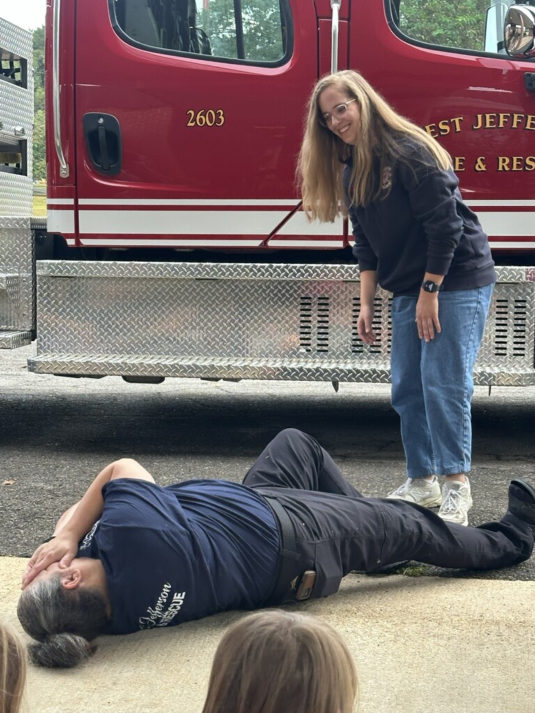 a lady fire fighter demonstrating "stop, drop, & roll" with another lady fire fighter looking on