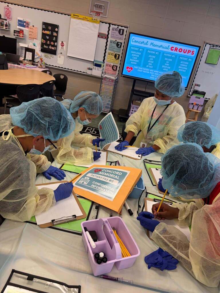 Students wearing hair nets and surgical gowns write on pieces of paper while sitting at tables.