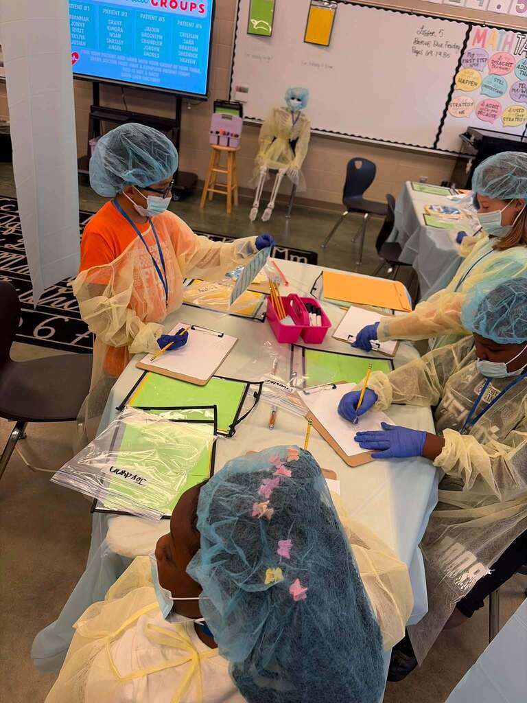 Students wearing hair nets and surgical gowns write on pieces of paper while sitting at tables.