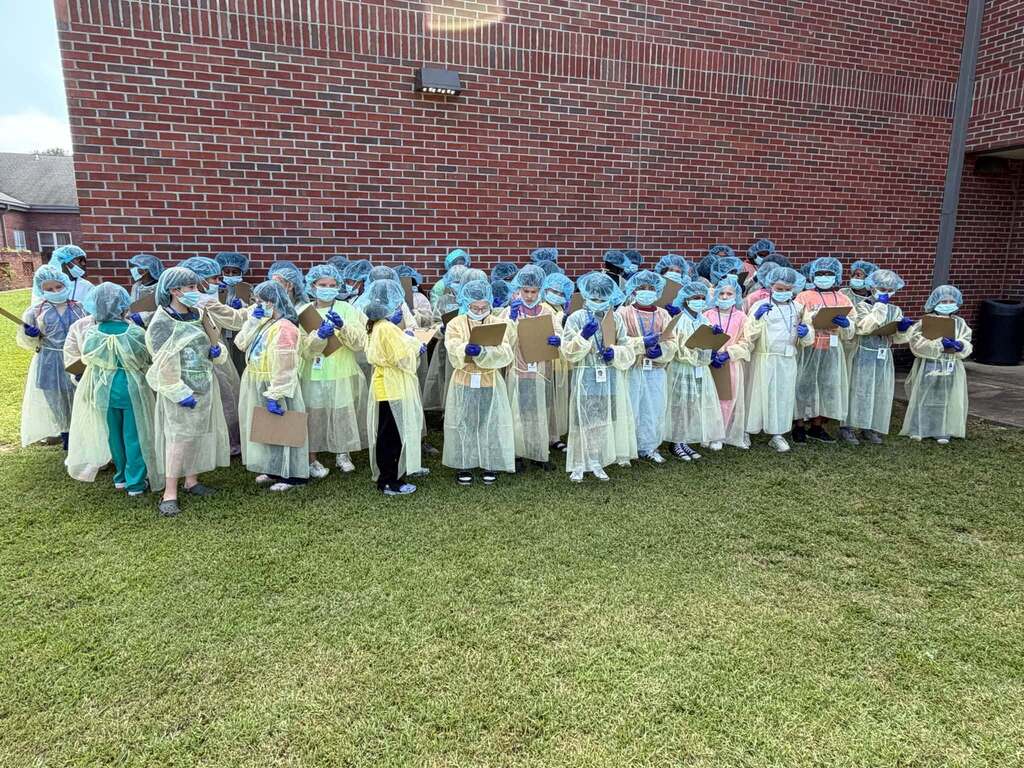 Students wearing hair nets and surgical gowns stand outside a school.
